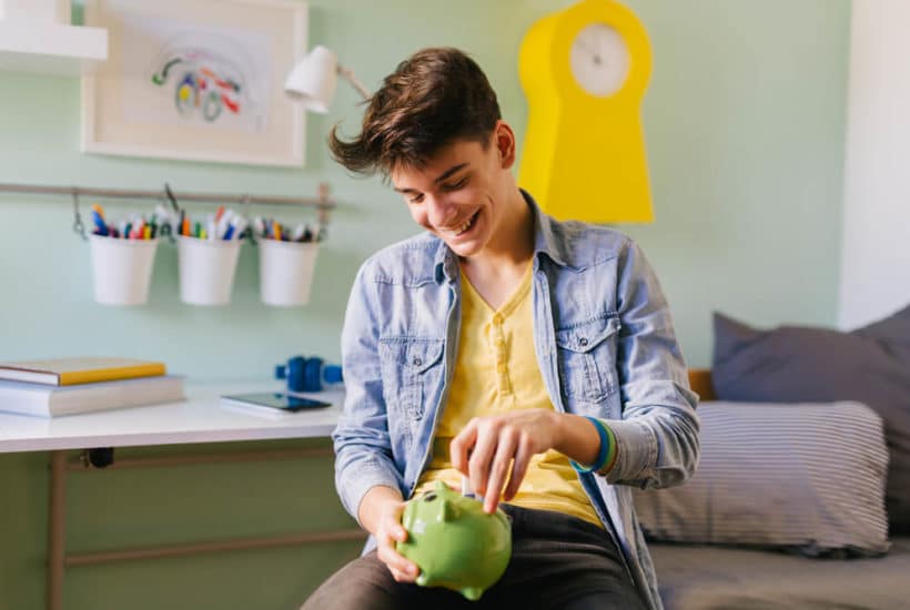 A teenage boy puts money into a green piggy bank for savings.