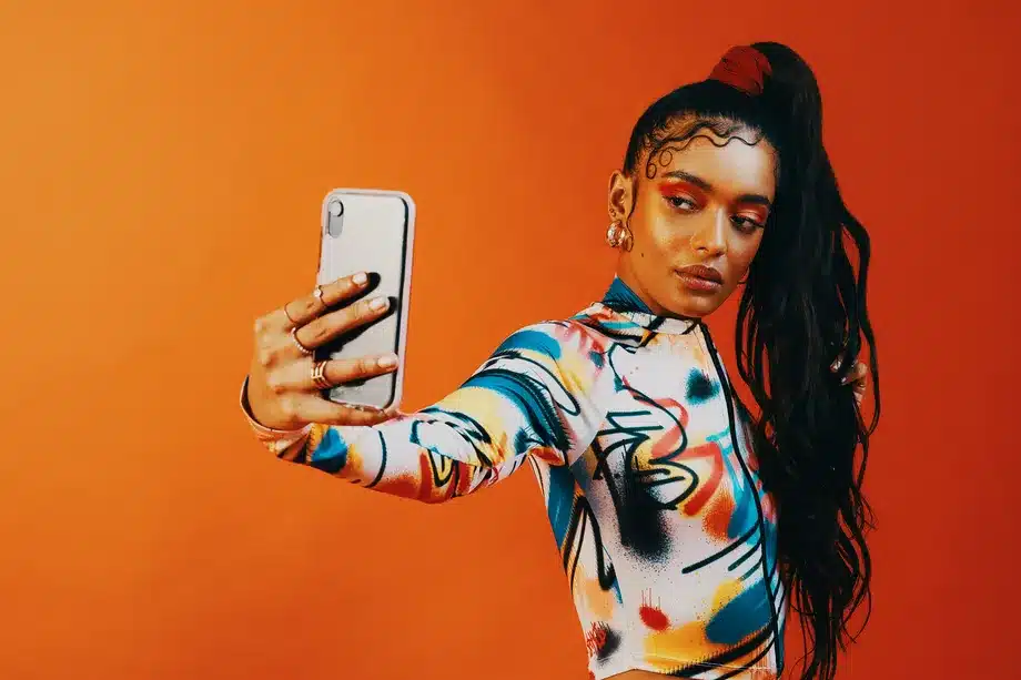 A female influencer takes a selfie wearing Gen Z fashion in front of an orange background.